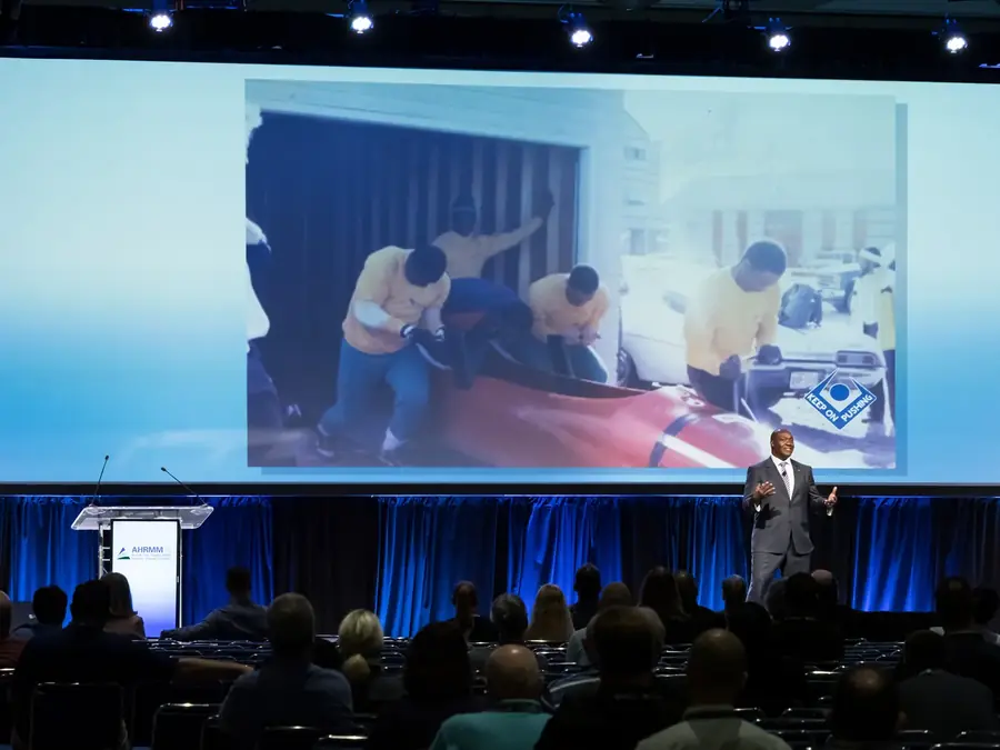 Devon Harris on a large conference stage with the Jamaican bobsled team on the screen behind him